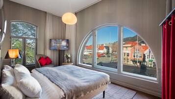 A modern hotel room with a large bed, a TV, and a view of a town through a large window.