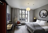 A hotel room with a bed, a desk, a chair, a large window, and a round mirror.