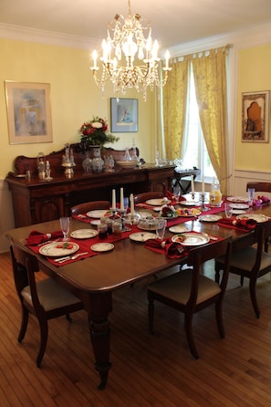 A dining room with a wooden table set for a meal, a chandelier, and a view of the dining area through a window with yellow curtains. - Auberge Chesley's Inn (Cornwall)