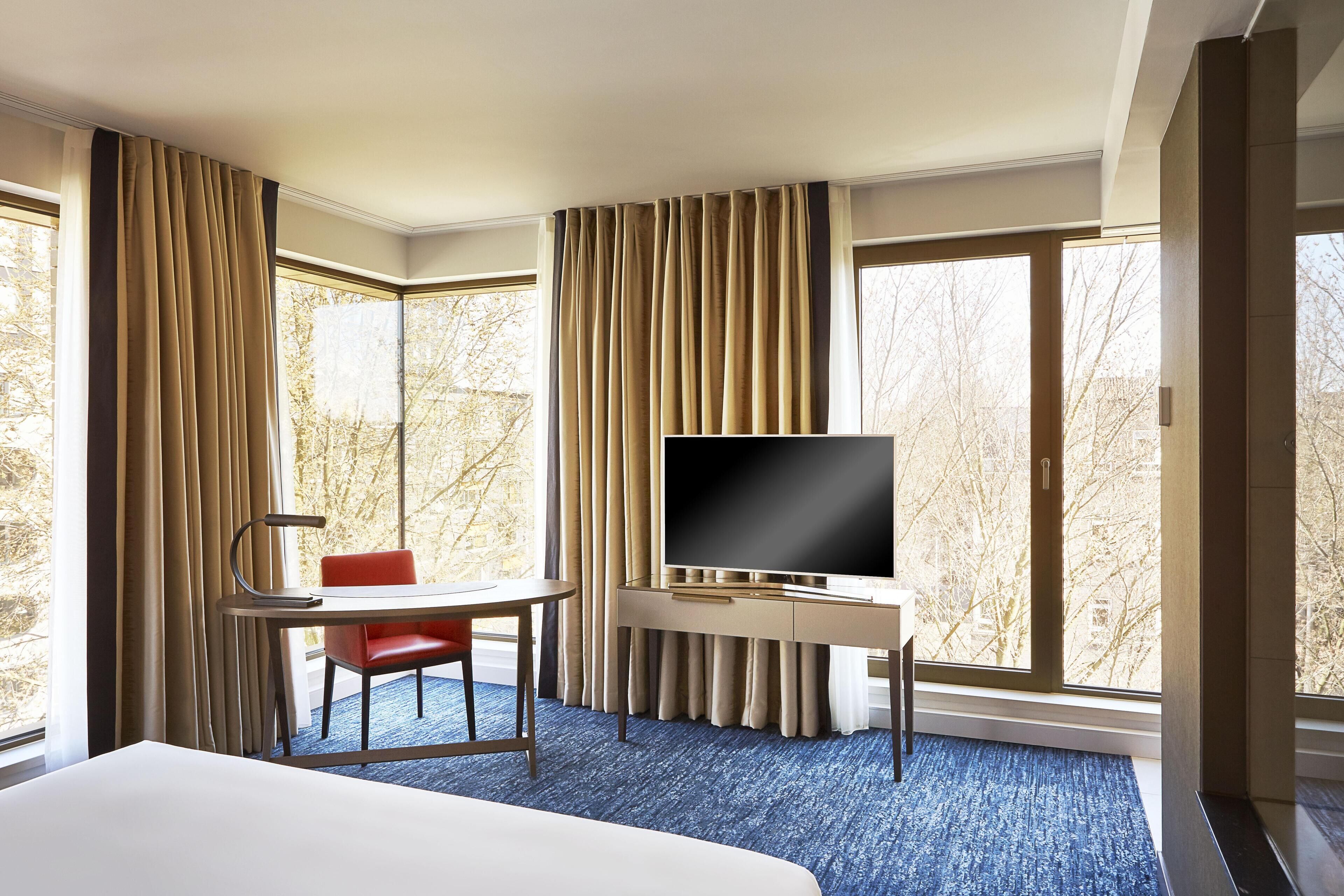 A hotel room with a bed, a desk with a TV, a chair, and large windows with curtains.
