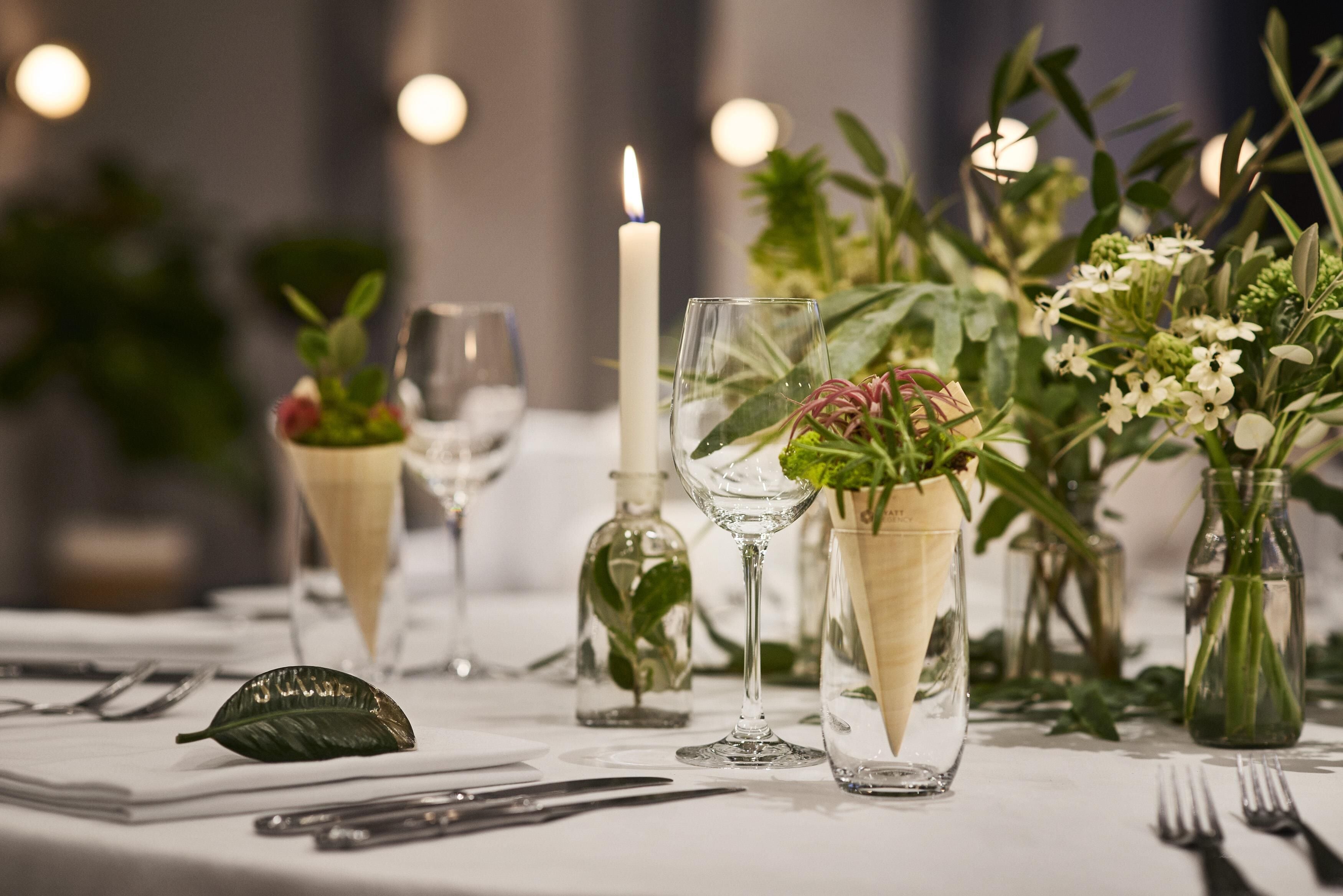 Ein gedeckter Tisch mit klaren Glasvasen, Blumen und Grünpflanzen, einer Kerze und einem gefalteten Servietten.