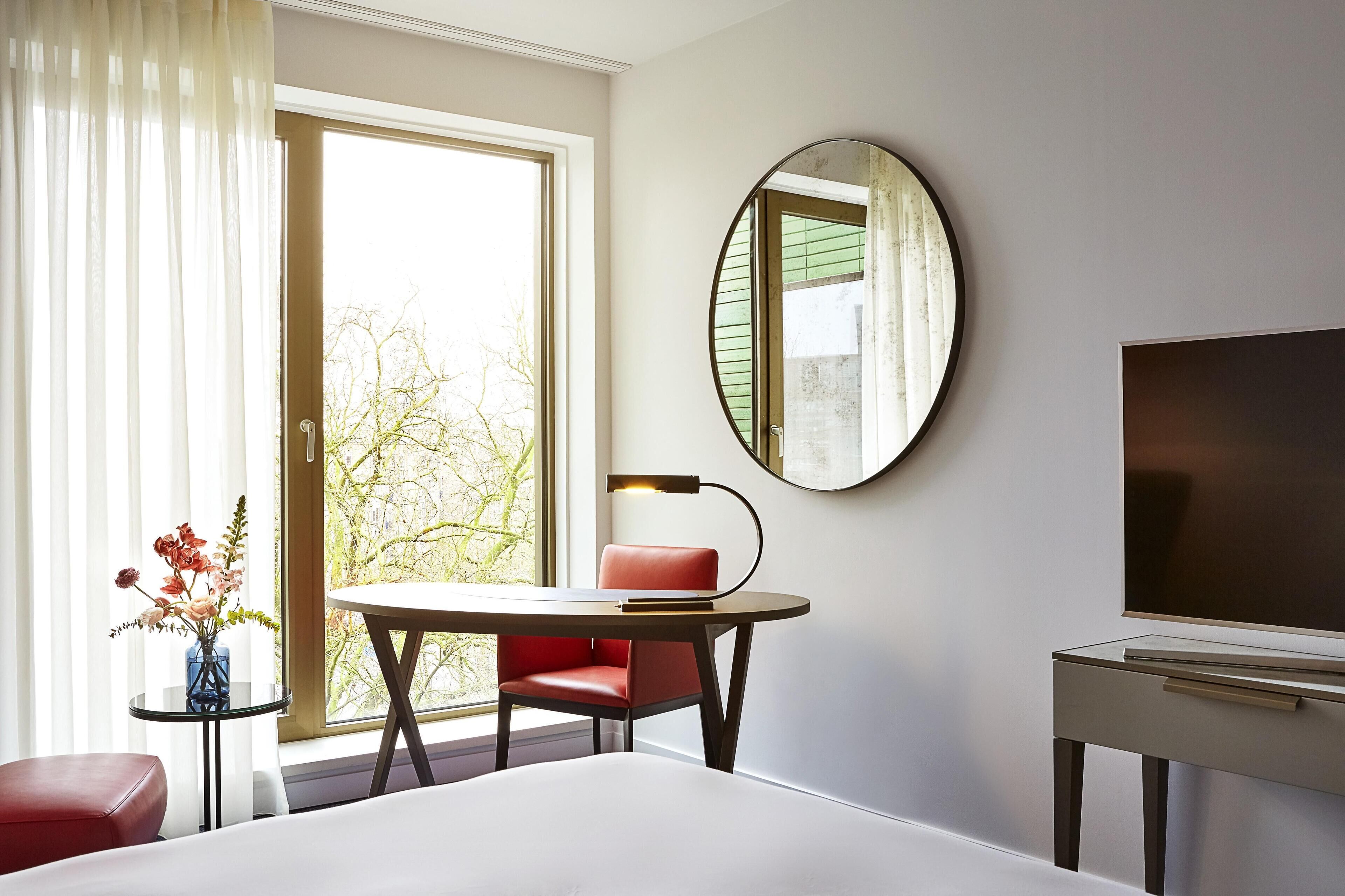 Una habitación de hotel con cama, escritorio con silla, televisión, espejo redondo y ventana con cortinas.