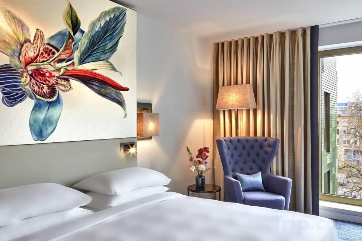 A hotel room with a bed, a blue armchair, a lamp, and a large floral painting on the wall.