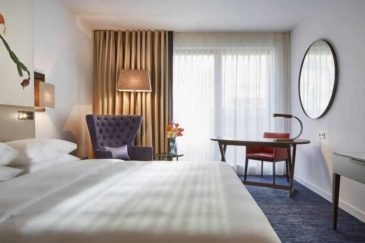 A hotel room with a large bed, a desk, a chair, a lamp, and a mirror.