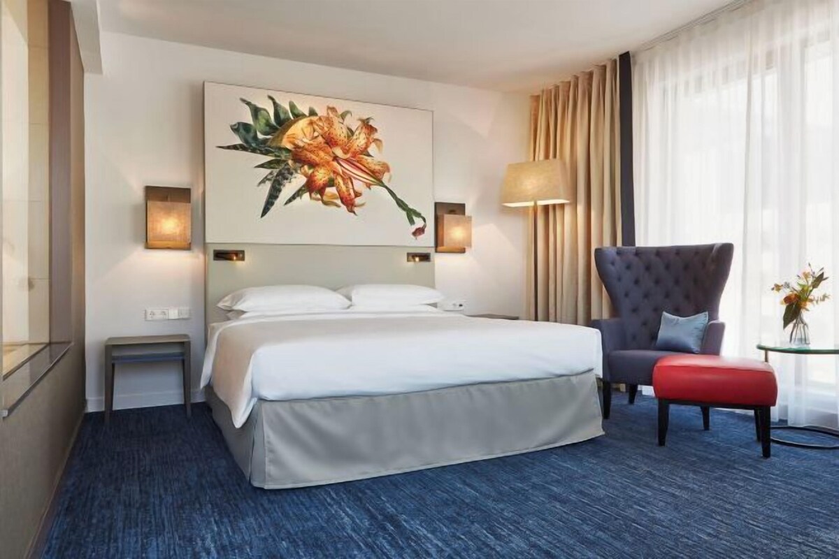 A hotel room with a large bed, a chair, a small table with a vase, and a wall-mounted painting.