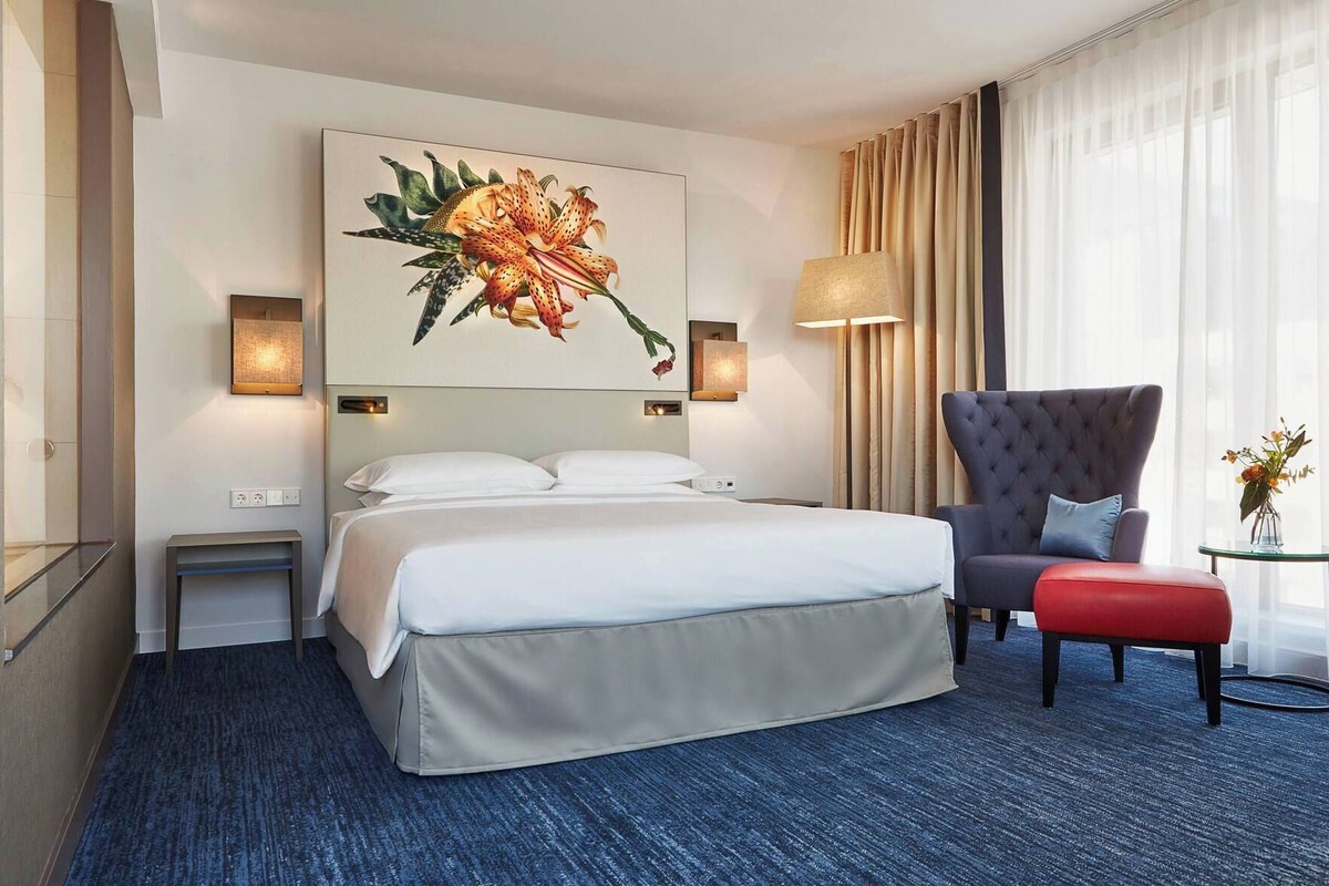 A hotel room with a large bed, a chair, a small table, and a wall-mounted painting.