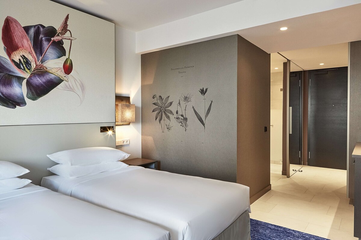 A hotel room with a large bed, a nightstand, a wall-mounted lamp, and a botanical print.