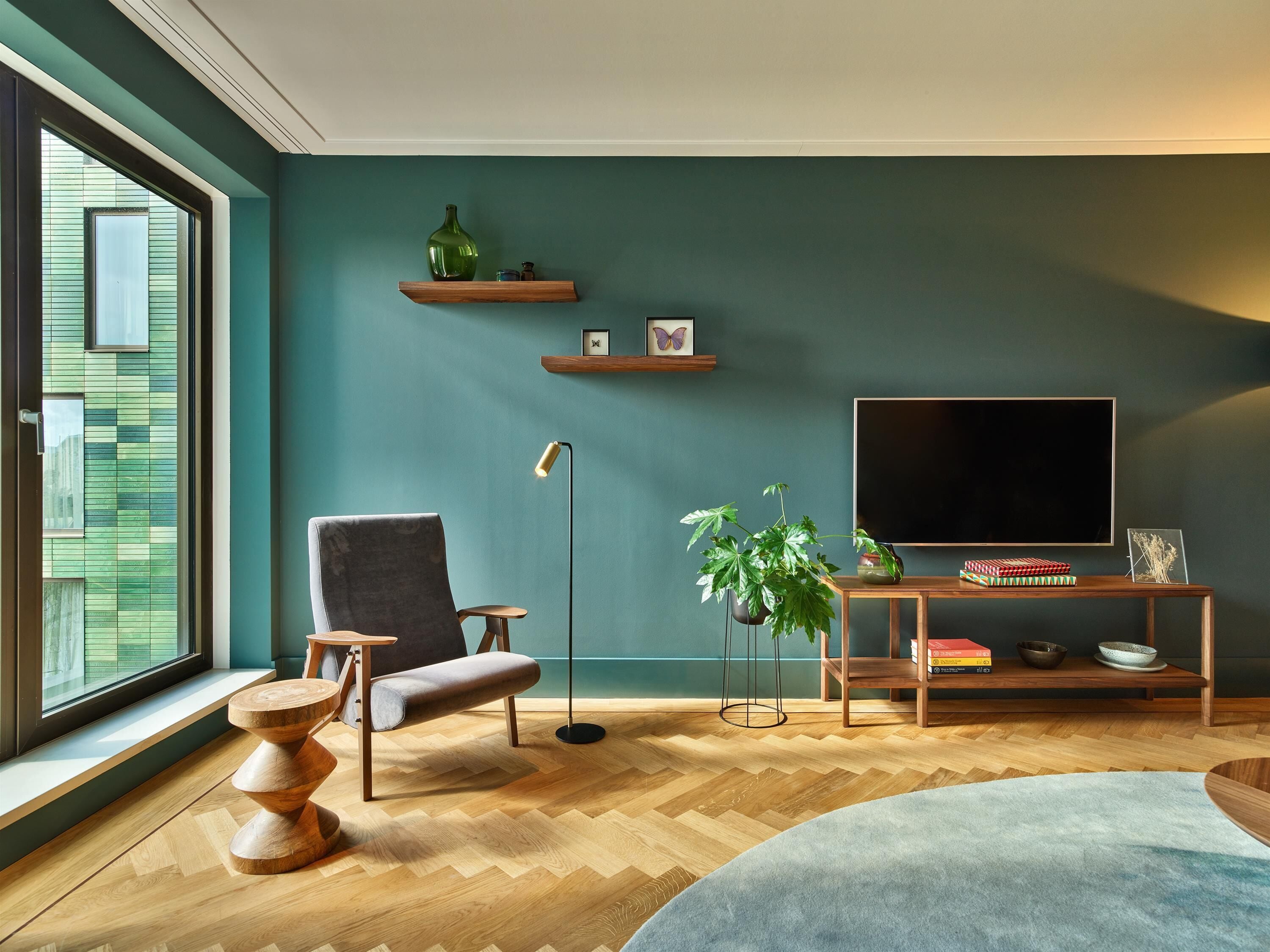 Un salon moderne avec une grande fenêtre, un fauteuil gris, une table basse en bois, un lampadaire, un téléviseur posé sur un meuble en bois et une plante en pot.