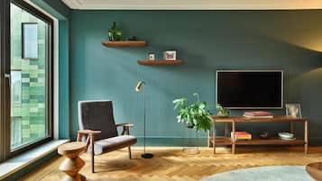 Un salon moderne avec une grande fenêtre, un fauteuil gris, une table basse en bois, un lampadaire, un téléviseur posé sur un meuble en bois et une plante en pot.