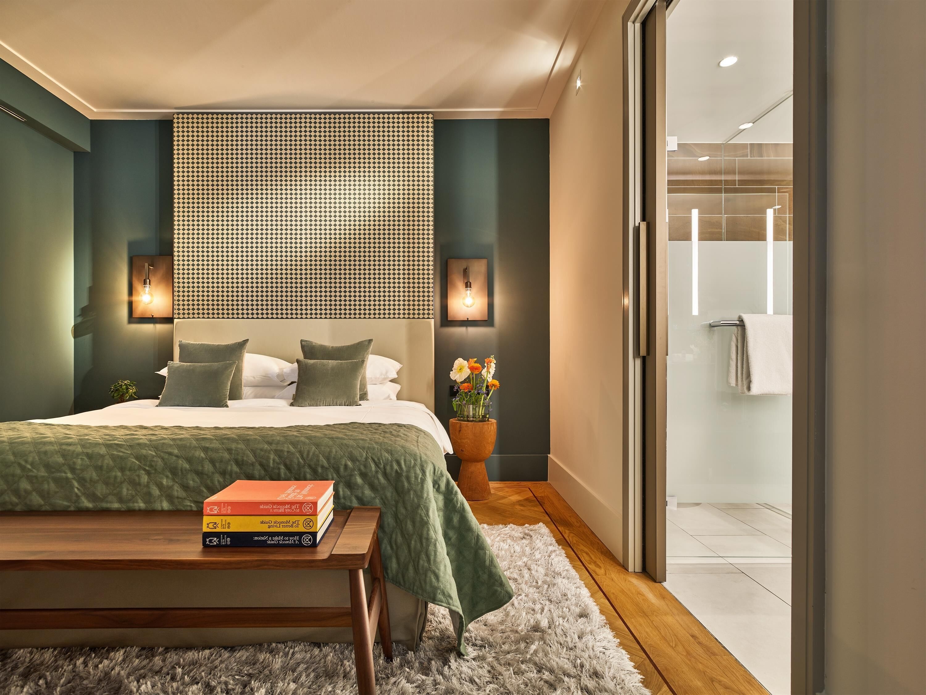 A modern bedroom with a large bed, bedside lamps, a wooden bedside table with books, and a view into the bathroom.