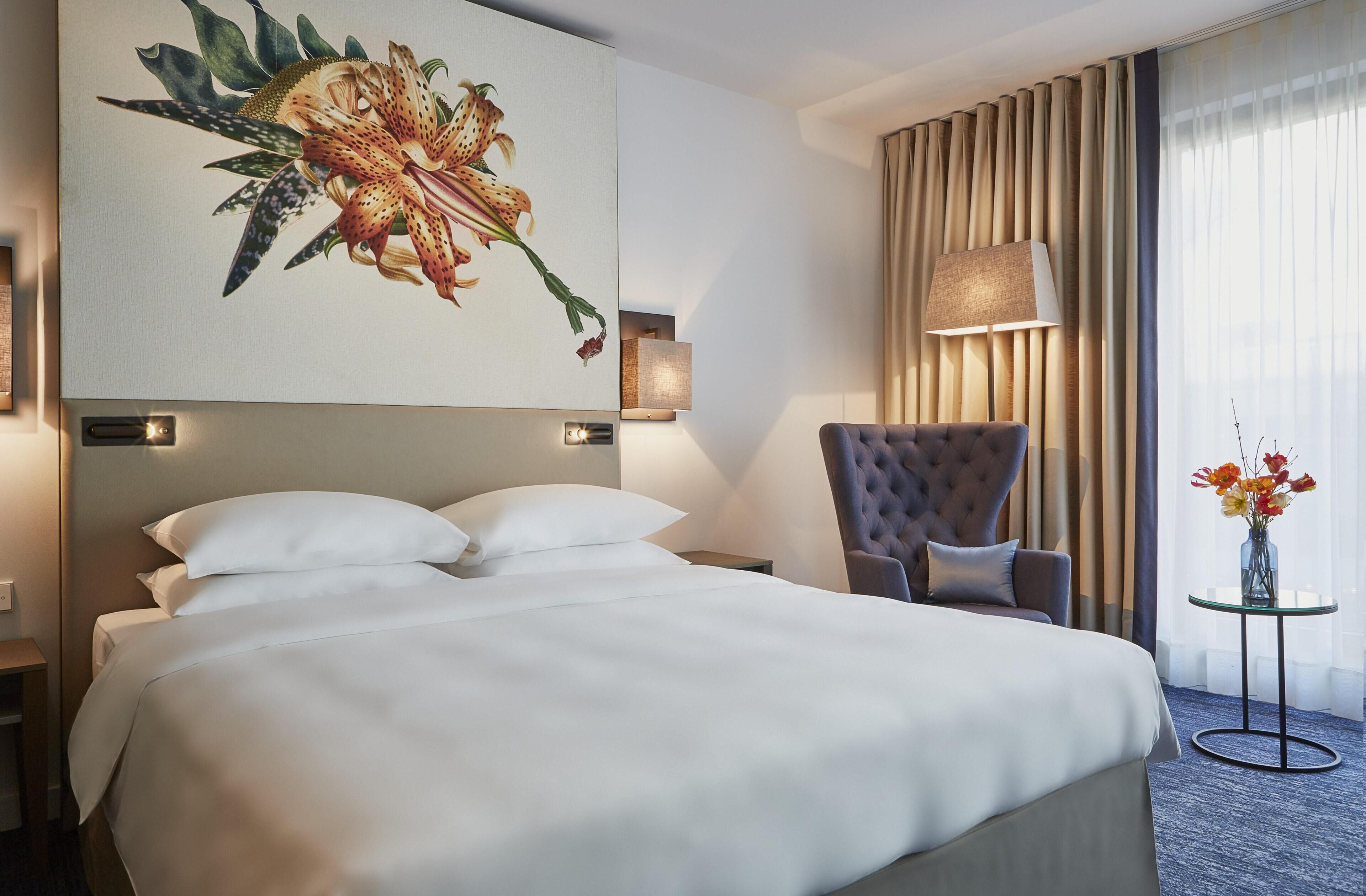Habitación de hotel con una cama grande, una silla, una lámpara y un cuadro de flores.