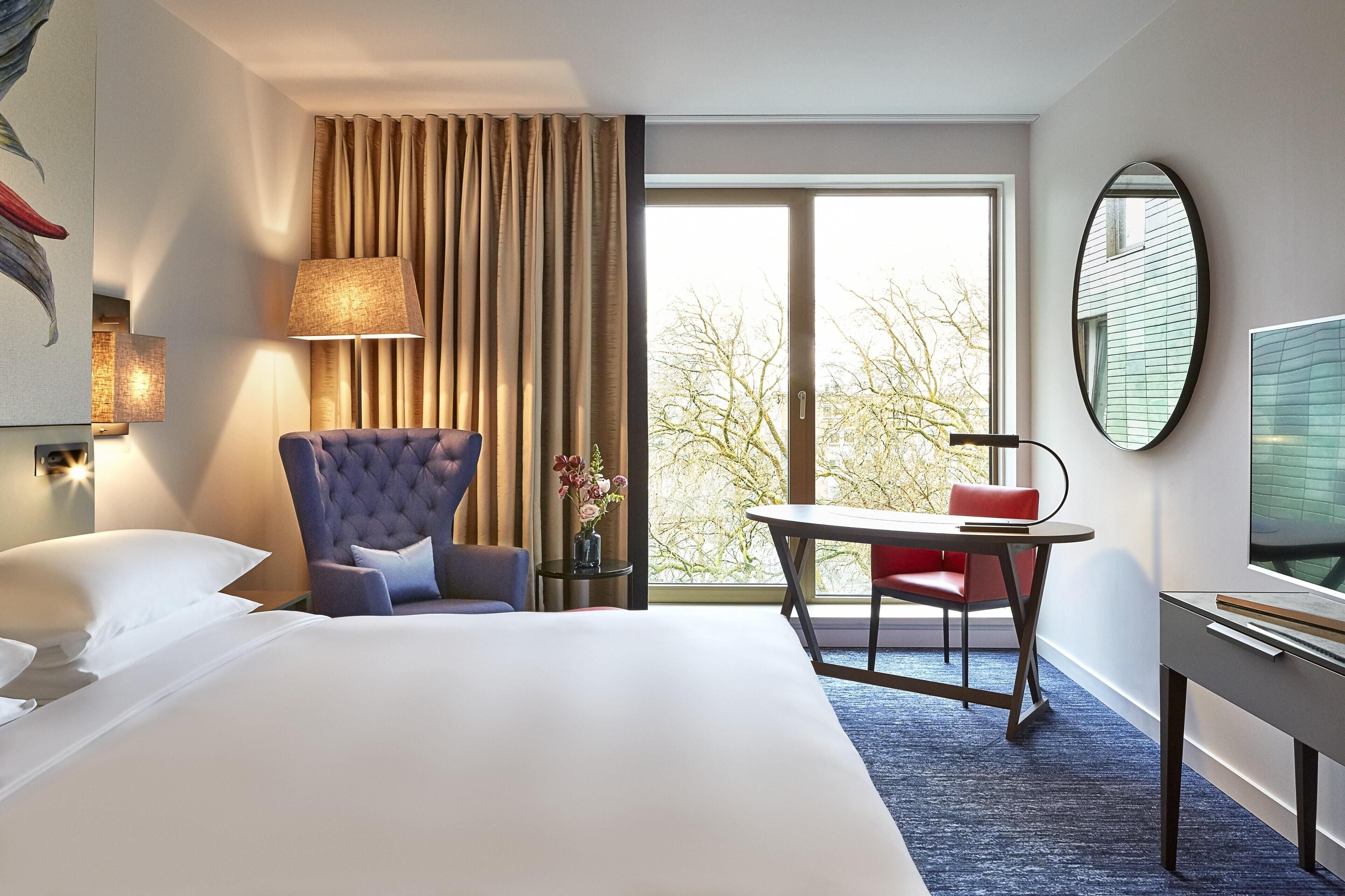 Una habitación de hotel con una cama grande, una zona de estar con una silla y una mesa, televisión y ventana con cortinas.