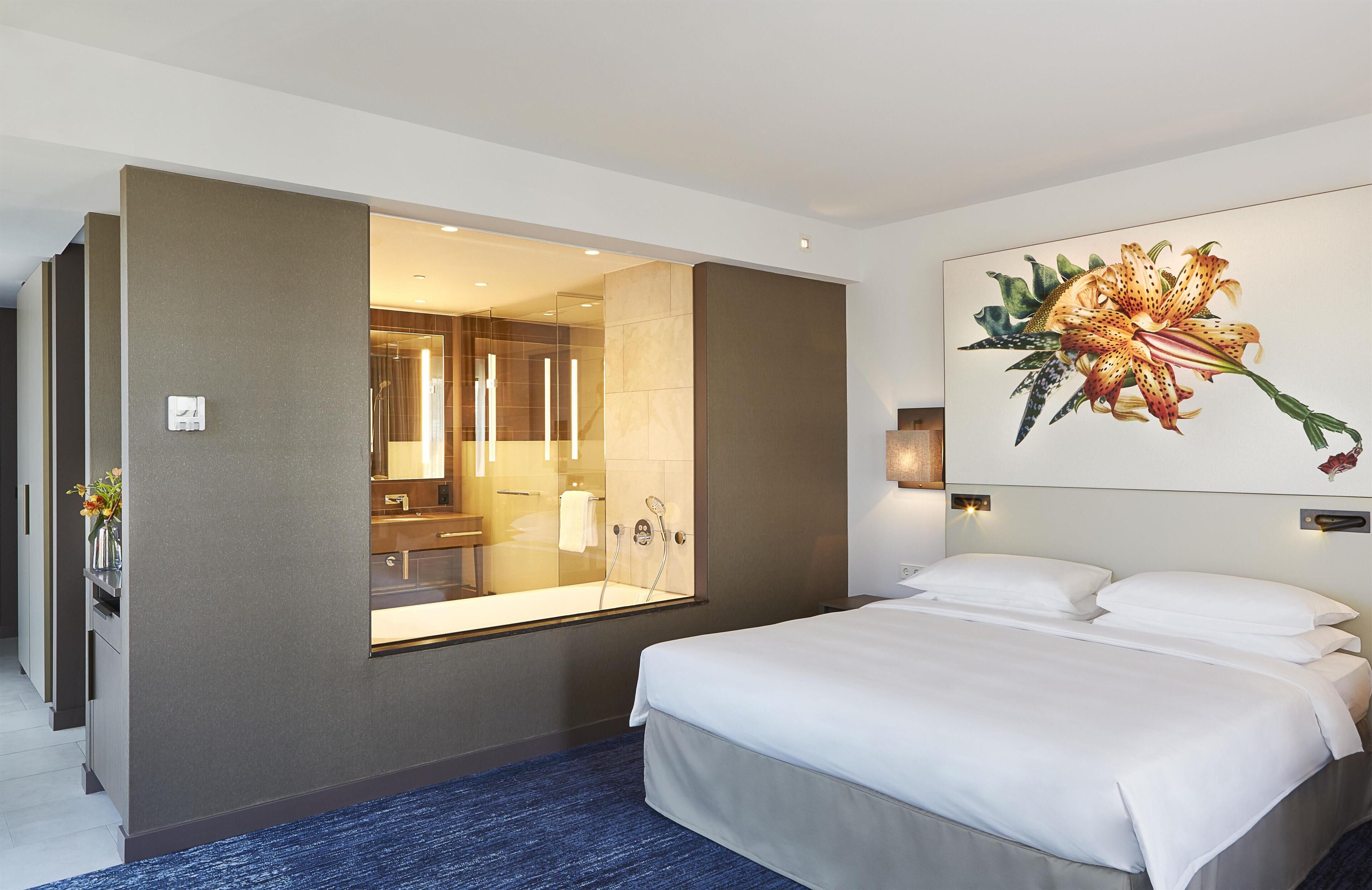 A hotel room with a large bed, a bathroom with a glass shower enclosure, and a wall-mounted painting of a flower.