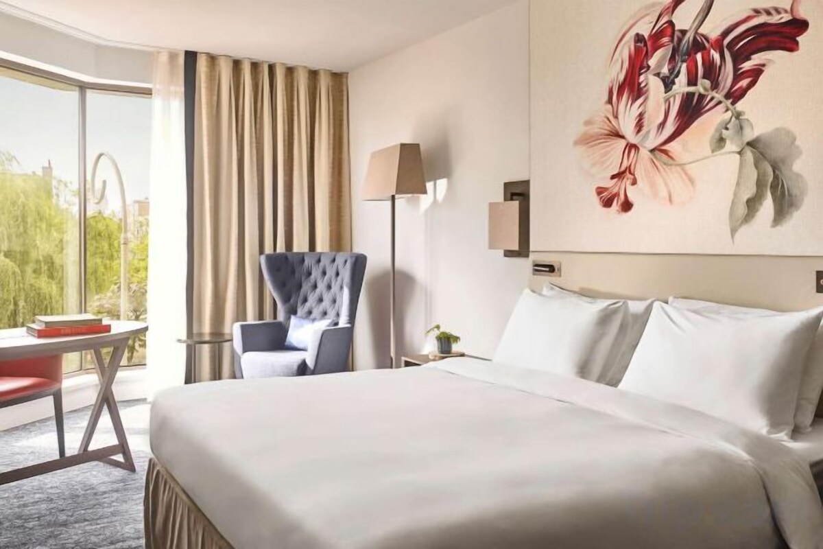 A hotel room with a large bed, a chair, a desk, and a window with curtains.
