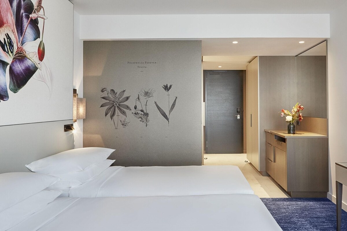 A modern hotel room with a large bed, a desk with a flat-screen TV, a small cabinet, and a wall-mounted artwork.