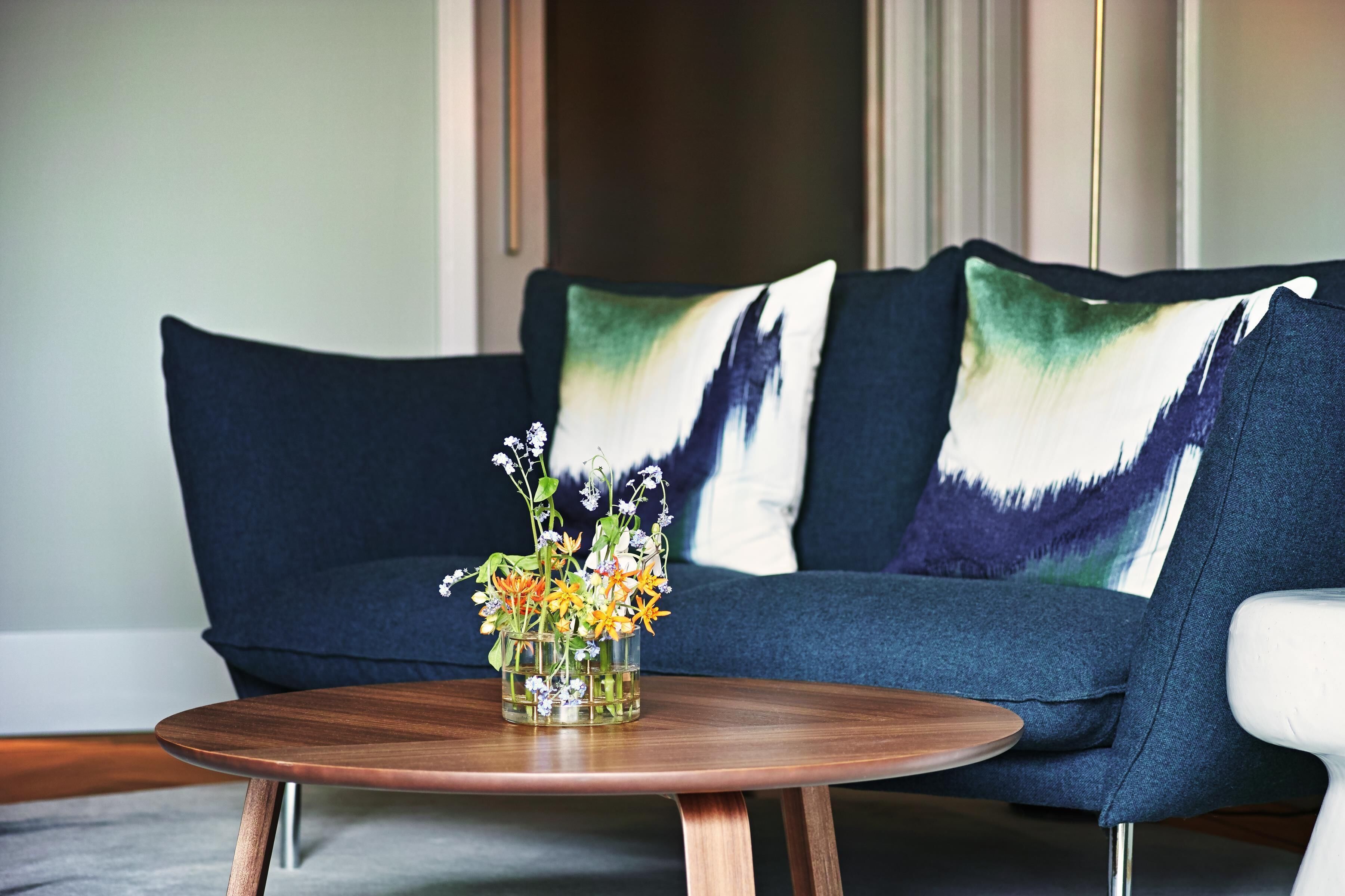 A modern living room with a dark blue sofa, two decorative pillows, and a wooden coffee table with a glass vase of flowers.