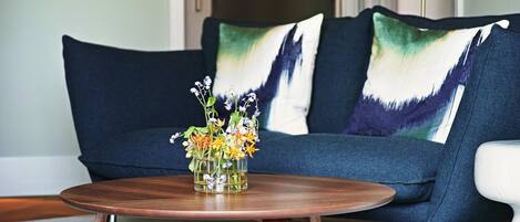 A modern living room with a dark blue sofa, two decorative pillows, and a wooden coffee table with a glass vase of flowers.