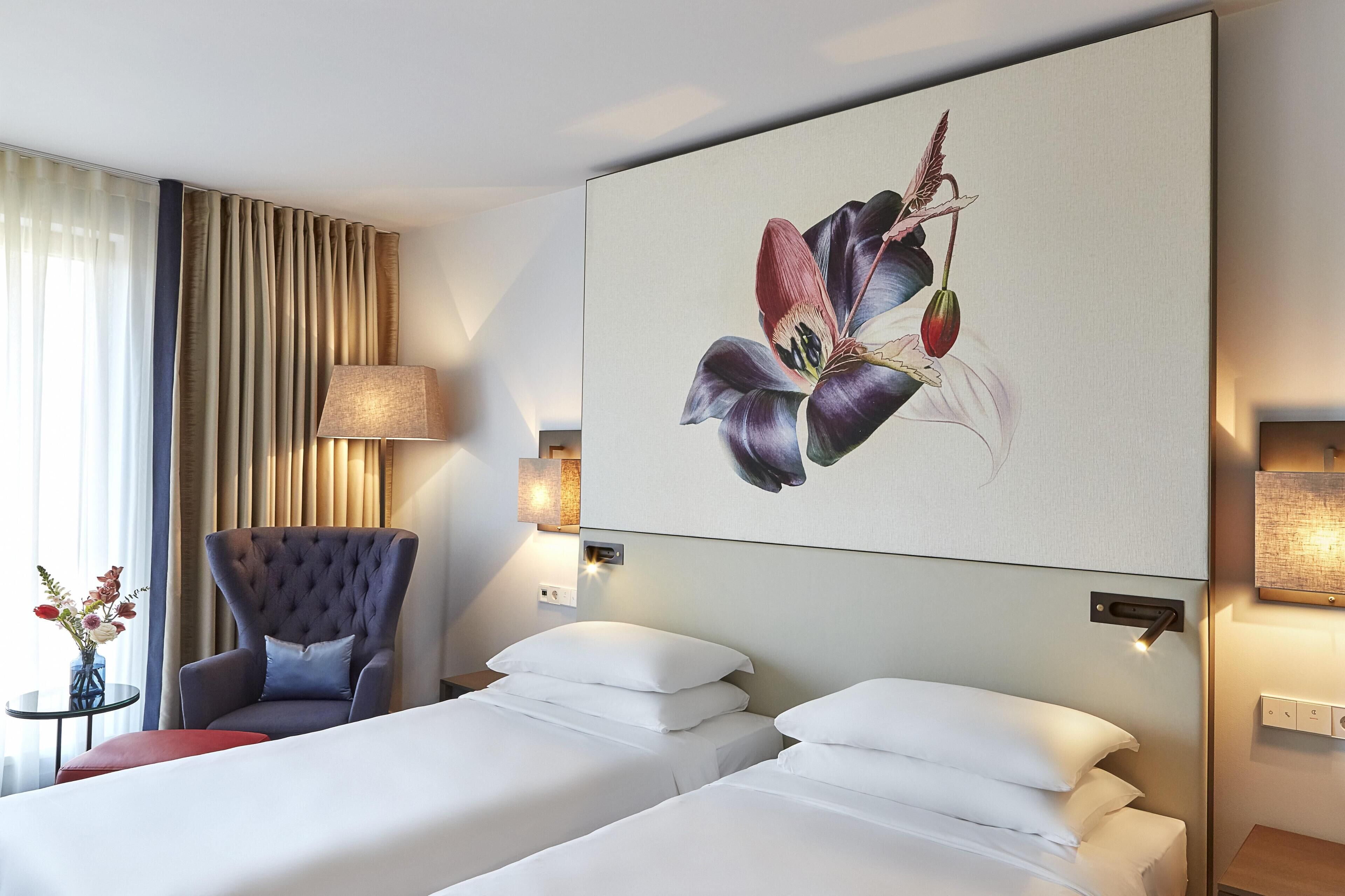 A hotel room with two beds, a chair, a lamp, and a wall-mounted artwork.