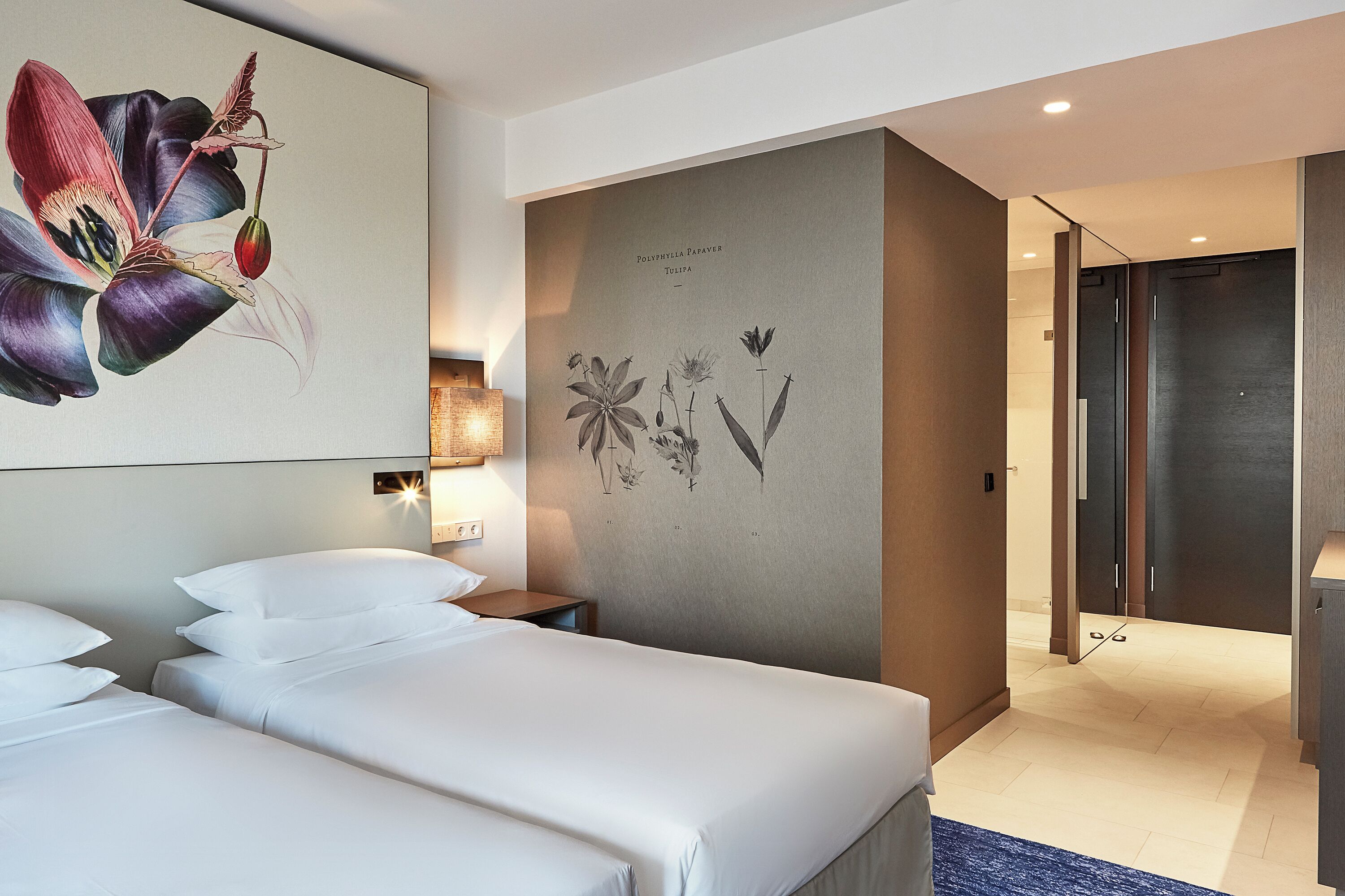Quarto de hotel com duas camas, uma ilustração botânica na parede e uma porta que dá para um corredor.