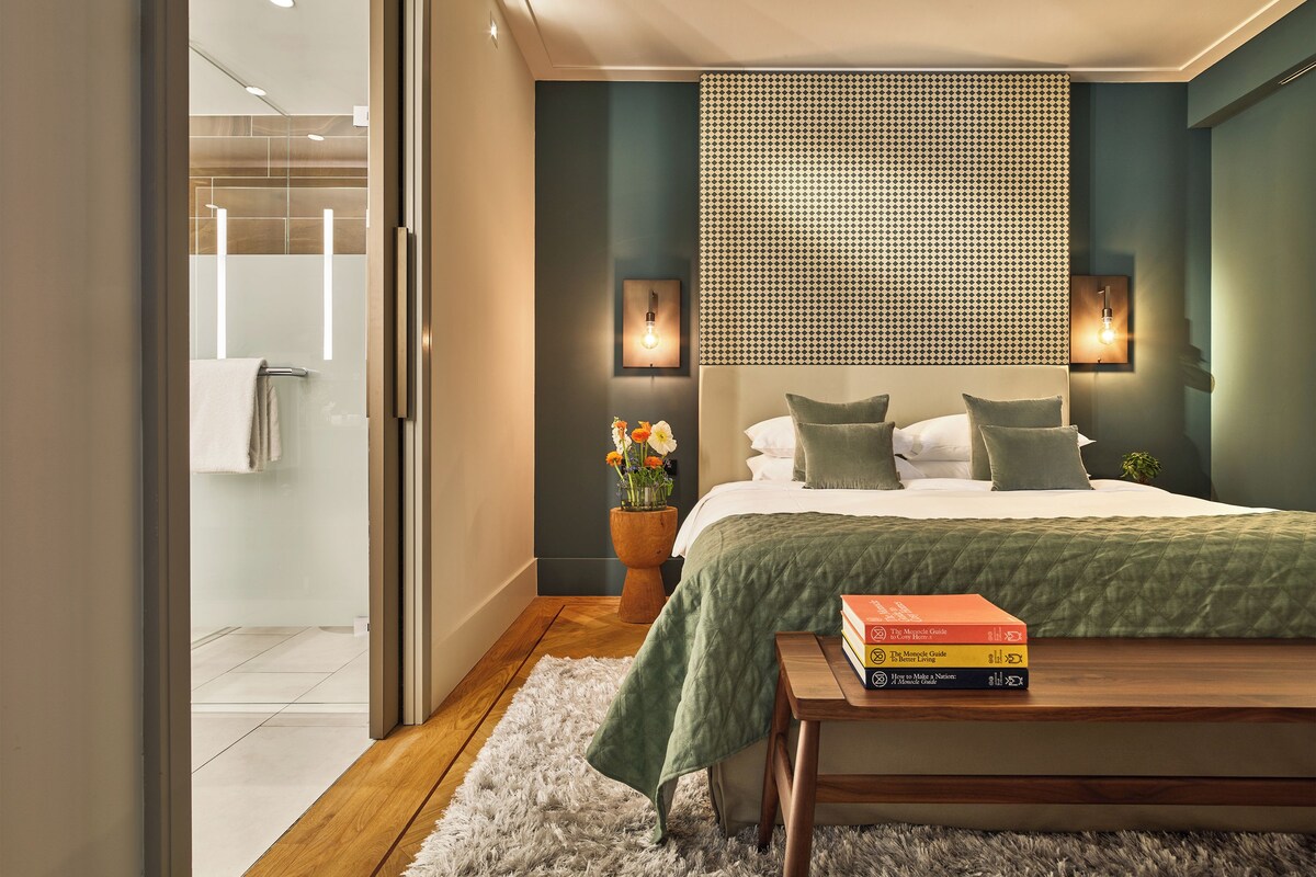 A modern bedroom with a large bed, a bedside table with books, and a patterned wall.