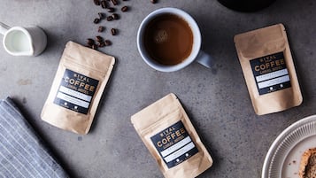 A coffee setup with a cup of coffee, two coffee bags, scattered coffee beans, a milk jug, and a coffee grinder on a textured surface.