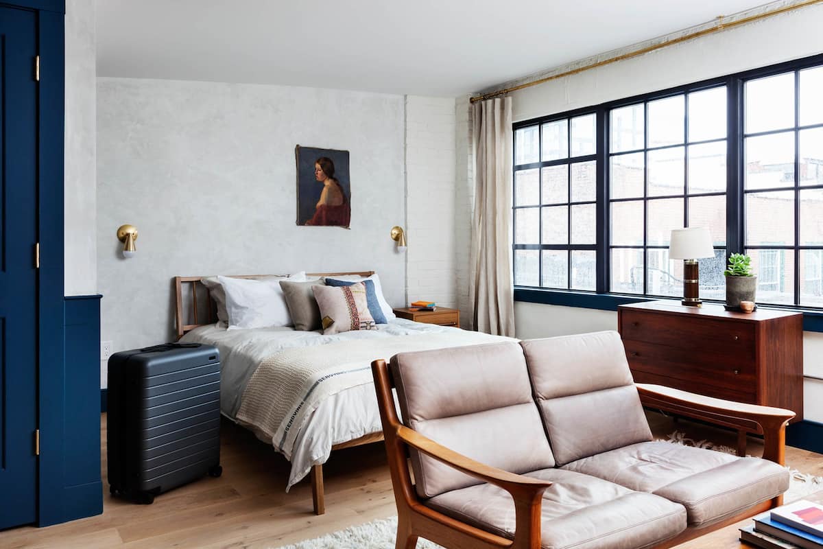 Une chambre à coucher avec un lit, une commode en bois, une chaise, une valise et une fenêtre avec des rideaux.
