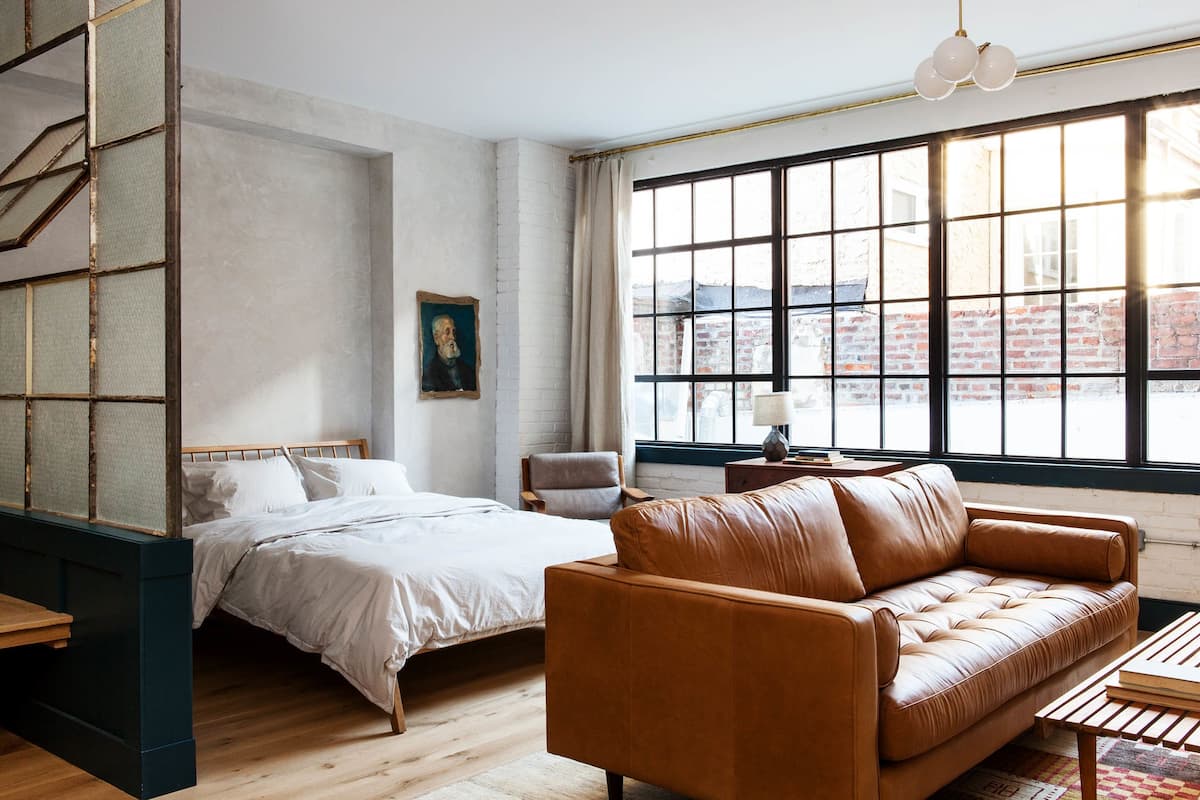 Une chambre à coucher comprenant un lit, un canapé en cuir, un bureau et une grande fenêtre donnant sur un immeuble en briques.