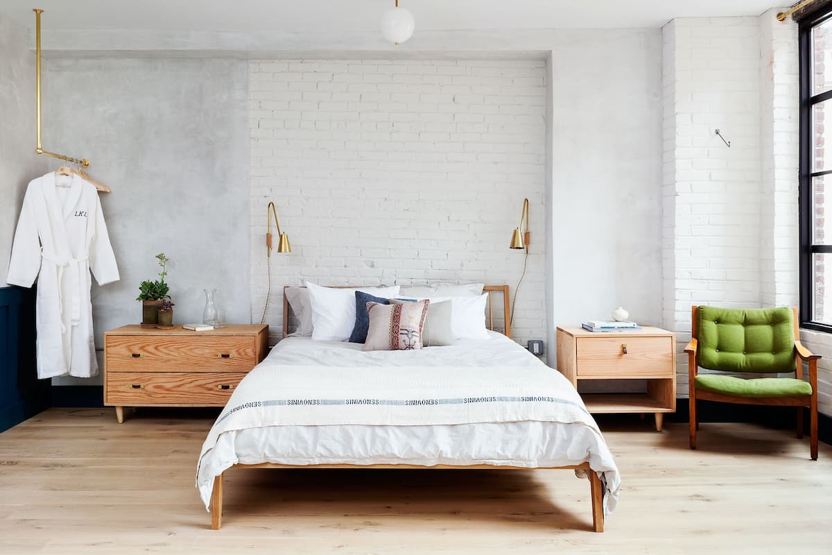Une chambre avec un mur en briques blanches, un lit en bois avec une literie blanche, deux tables de chevet avec des lampes, un fauteuil vert et un peignoir suspendu.