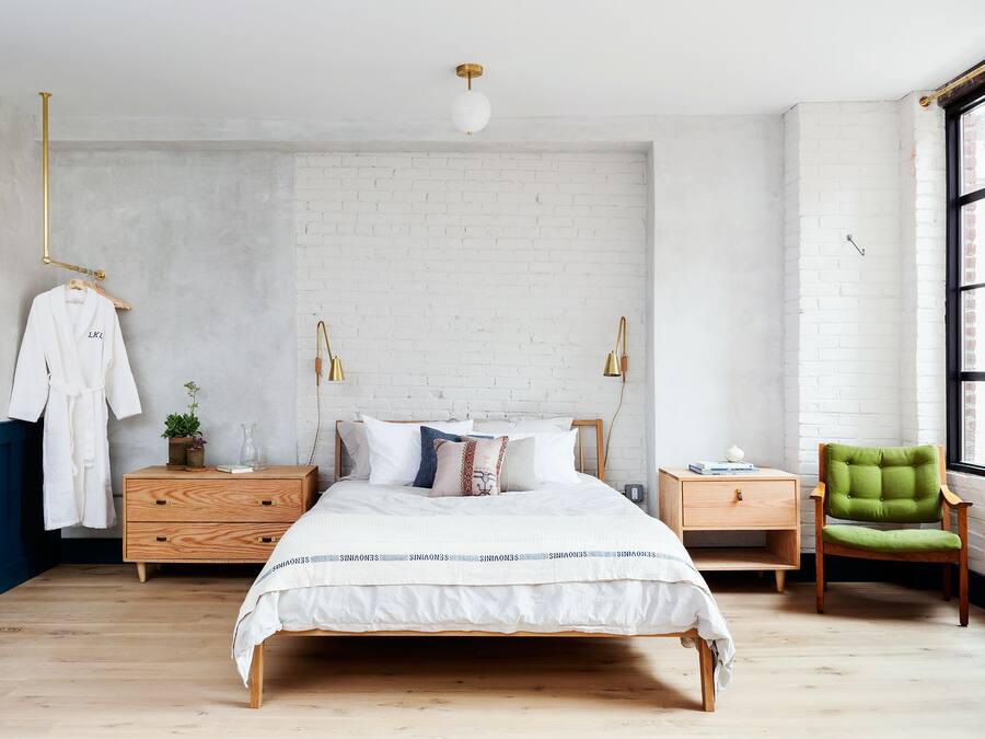 Un dormitorio con pared de ladrillo blanco, una cama de madera con ropa blanca, dos mesitas de noche con lámparas, una silla verde y una bata colgada.