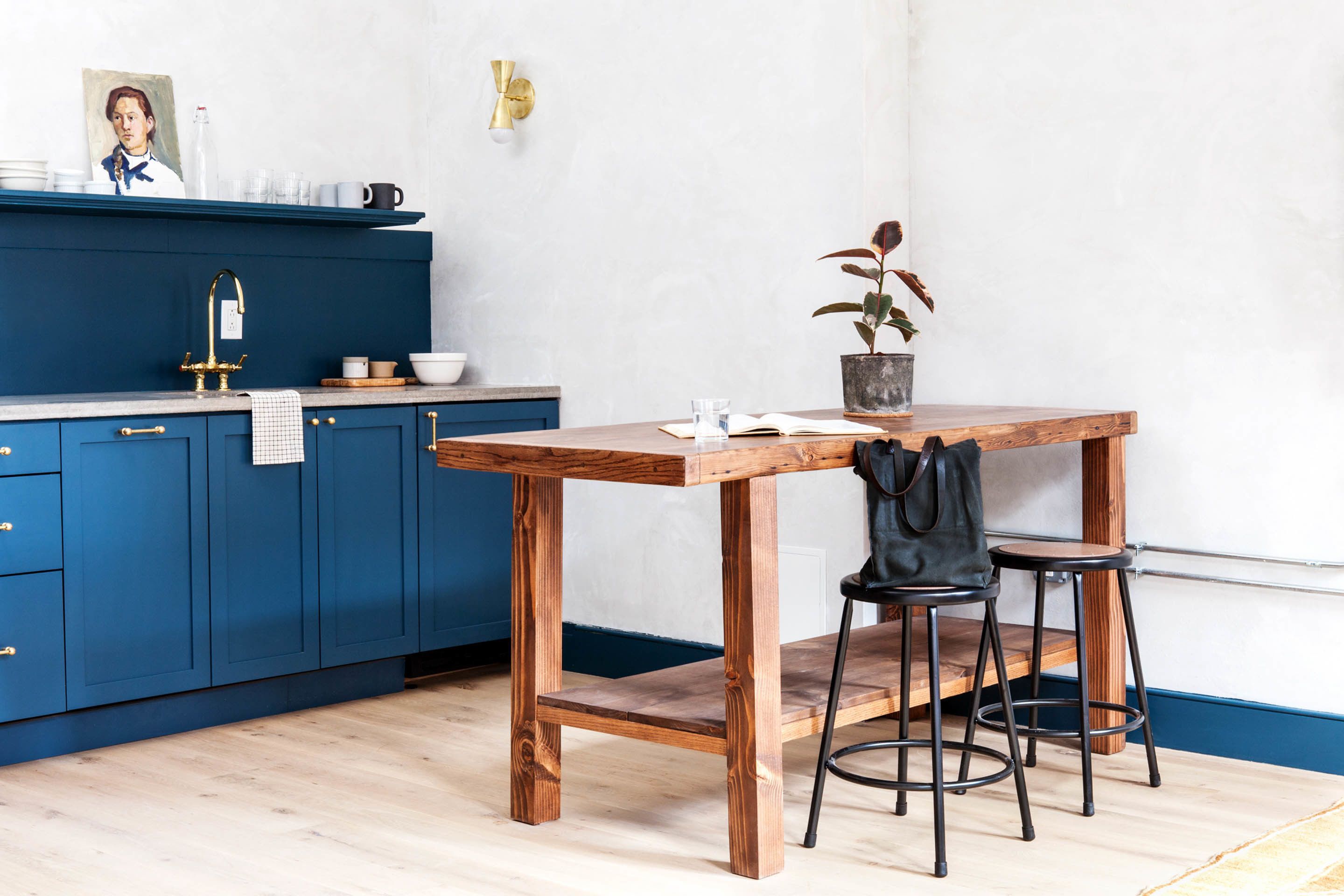 A kitchen with blue cabinets, a wooden table, two stools, a potted plant, and a wall-mounted light fixture.