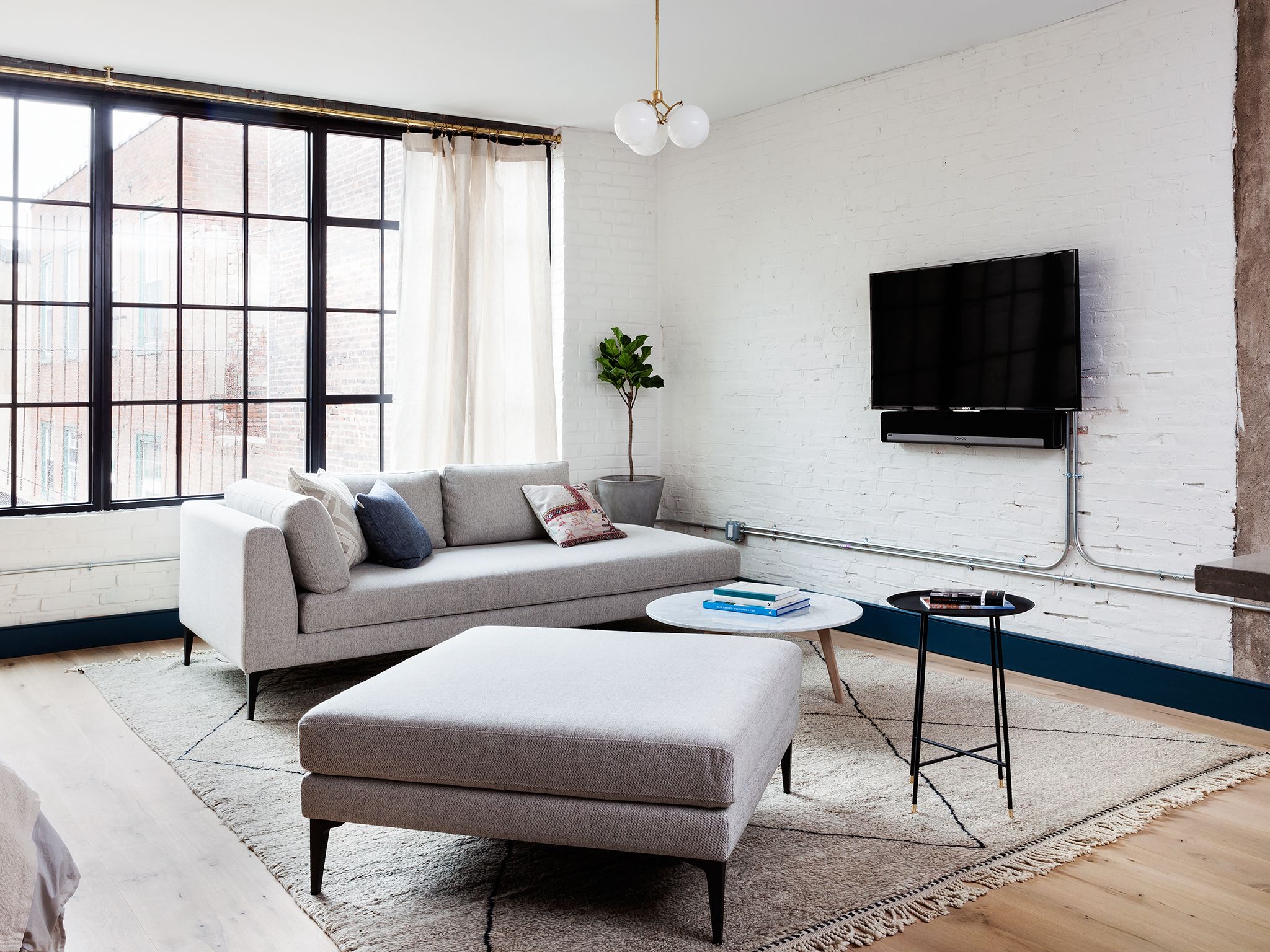 Un salon moderne avec un canapé, un pouf et une table basse ronde. Un téléviseur à écran plat est fixé au mur, et on peut voir une plante en pot.