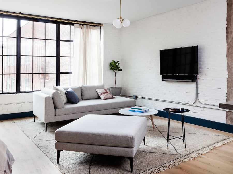 Un salon moderne avec un canapé, un pouf et une table basse ronde. Un téléviseur à écran plat est fixé au mur, et on peut voir une plante en pot.