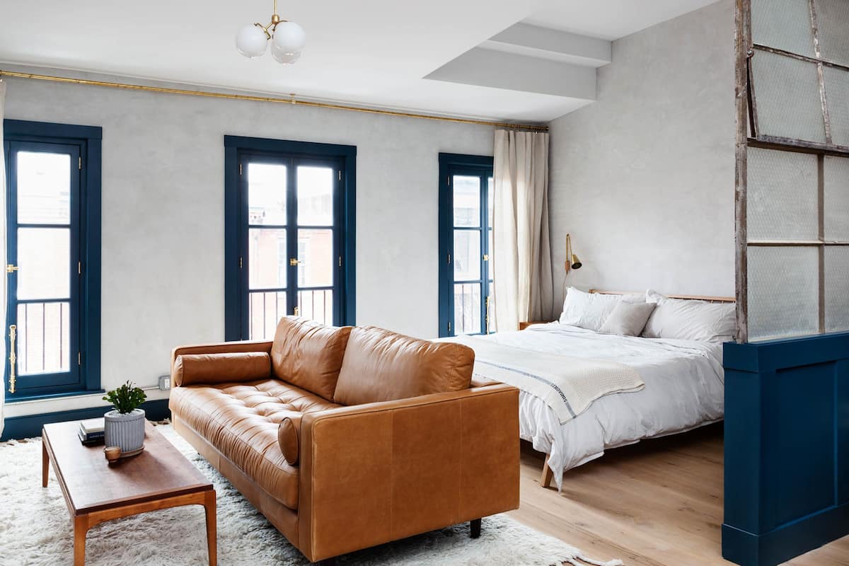 Une chambre moderne avec un canapé en cuir marron, une table basse en bois, un lit blanc et de grandes fenêtres aux montures bleues.