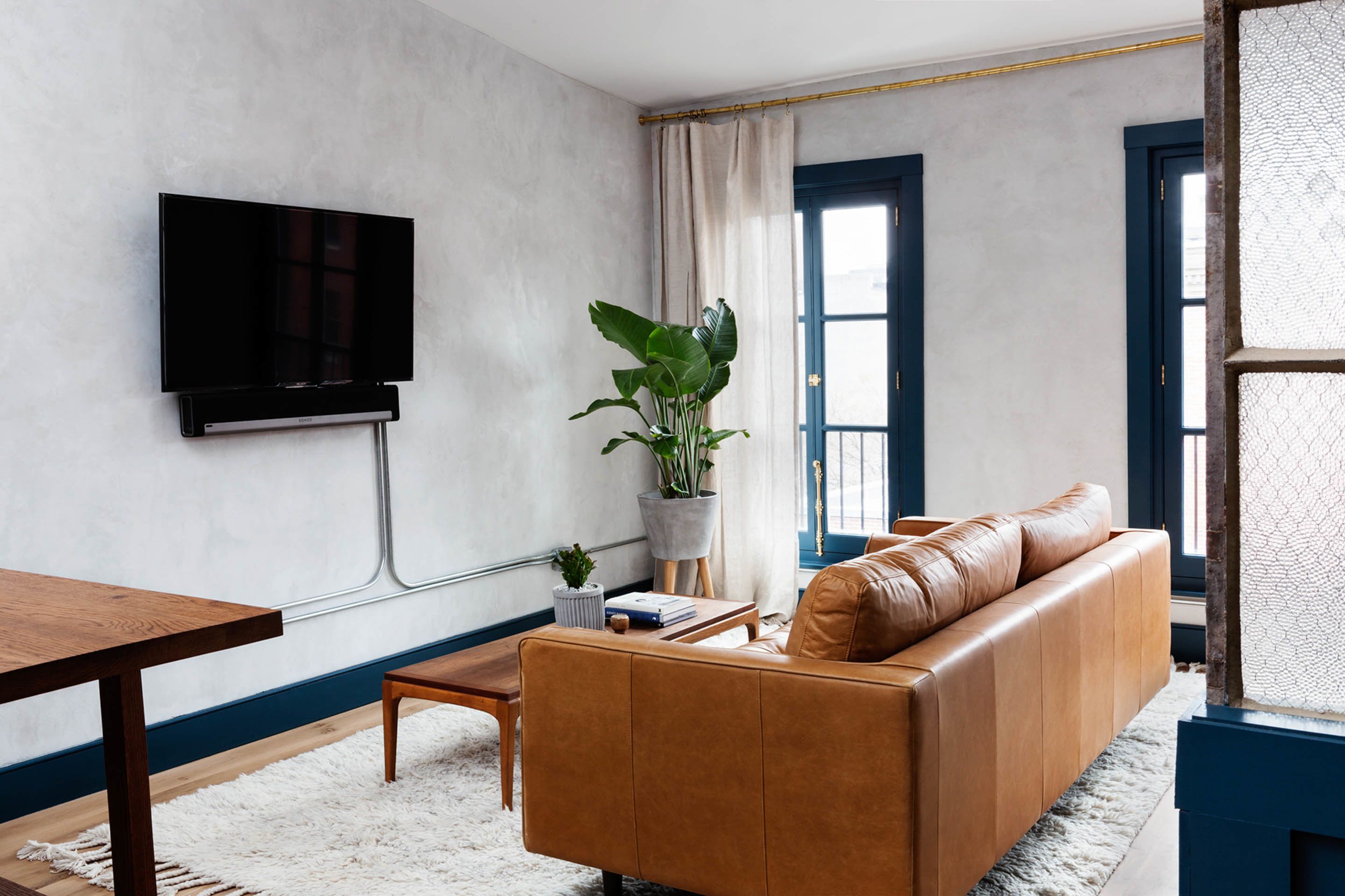 Una sala de estar moderna con un sofá de cuero, una mesa de centro, un televisor de pantalla plana montado en la pared, una planta en maceta y un gran ventanal con cortinas.
