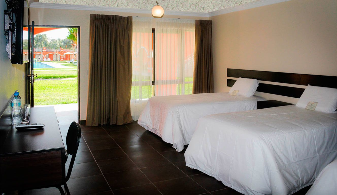 A hotel room with two beds, a desk with a chair, a window with curtains, and a view of a swimming pool.