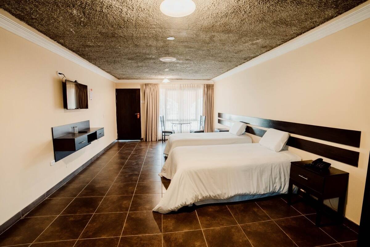 Habitación de hotel con dos camas, un escritorio pequeño, una silla, un televisor y un baño con espejo y lavamanos.