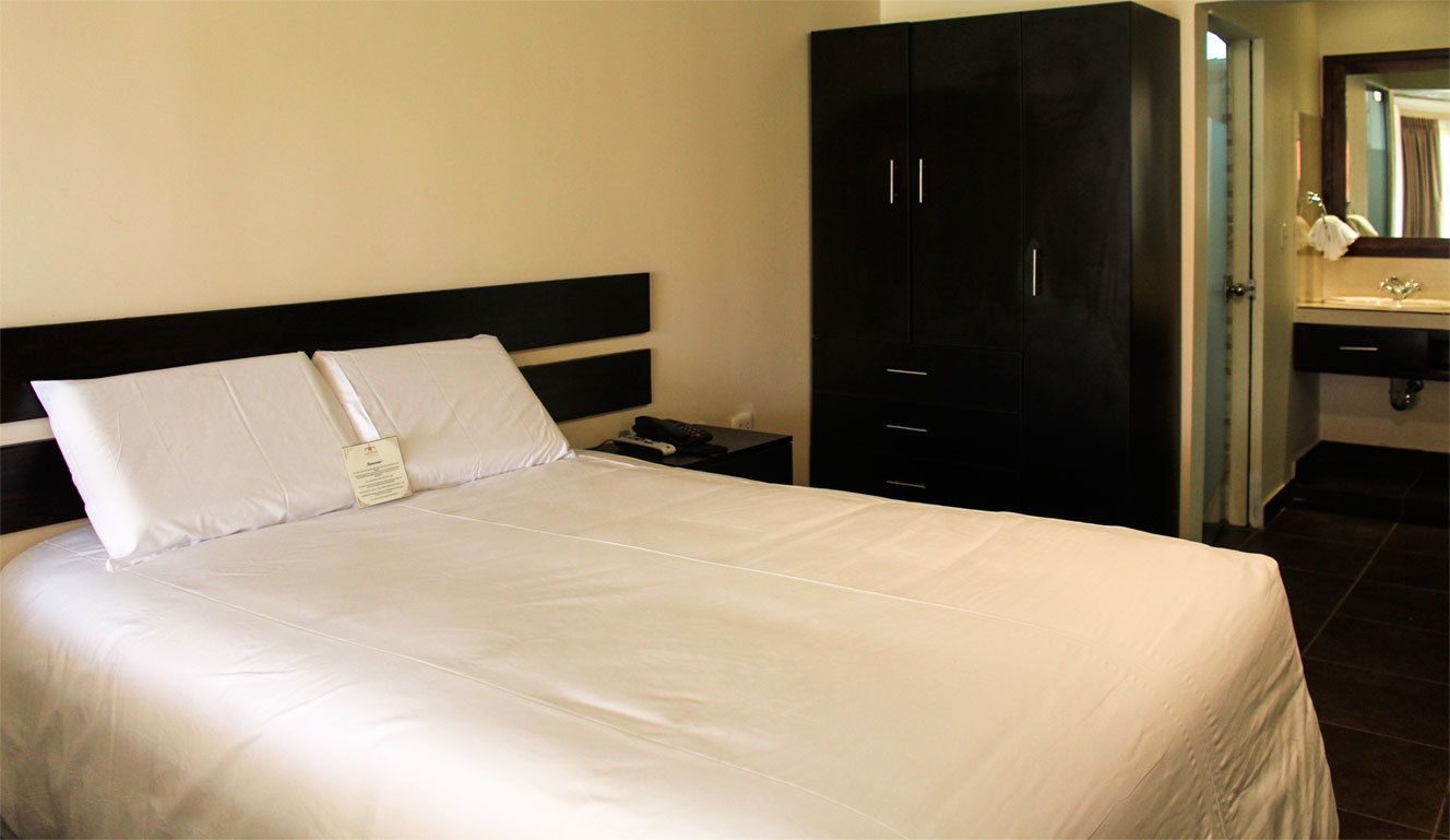 A hotel room with a bed, a nightstand, a black wardrobe, and a bathroom with a sink and mirror.