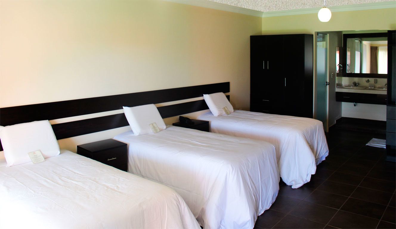 Habitación de hotel con dos camas individuales, un armario de madera oscura, un baño con espejo y lavamanos, y una lámpara en el techo.