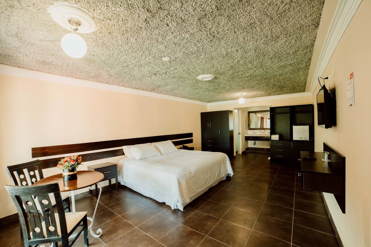 Habitación de hotel con cama, escritorio con silla, una mesita con flores, televisor y baño con espejo y lavamanos.