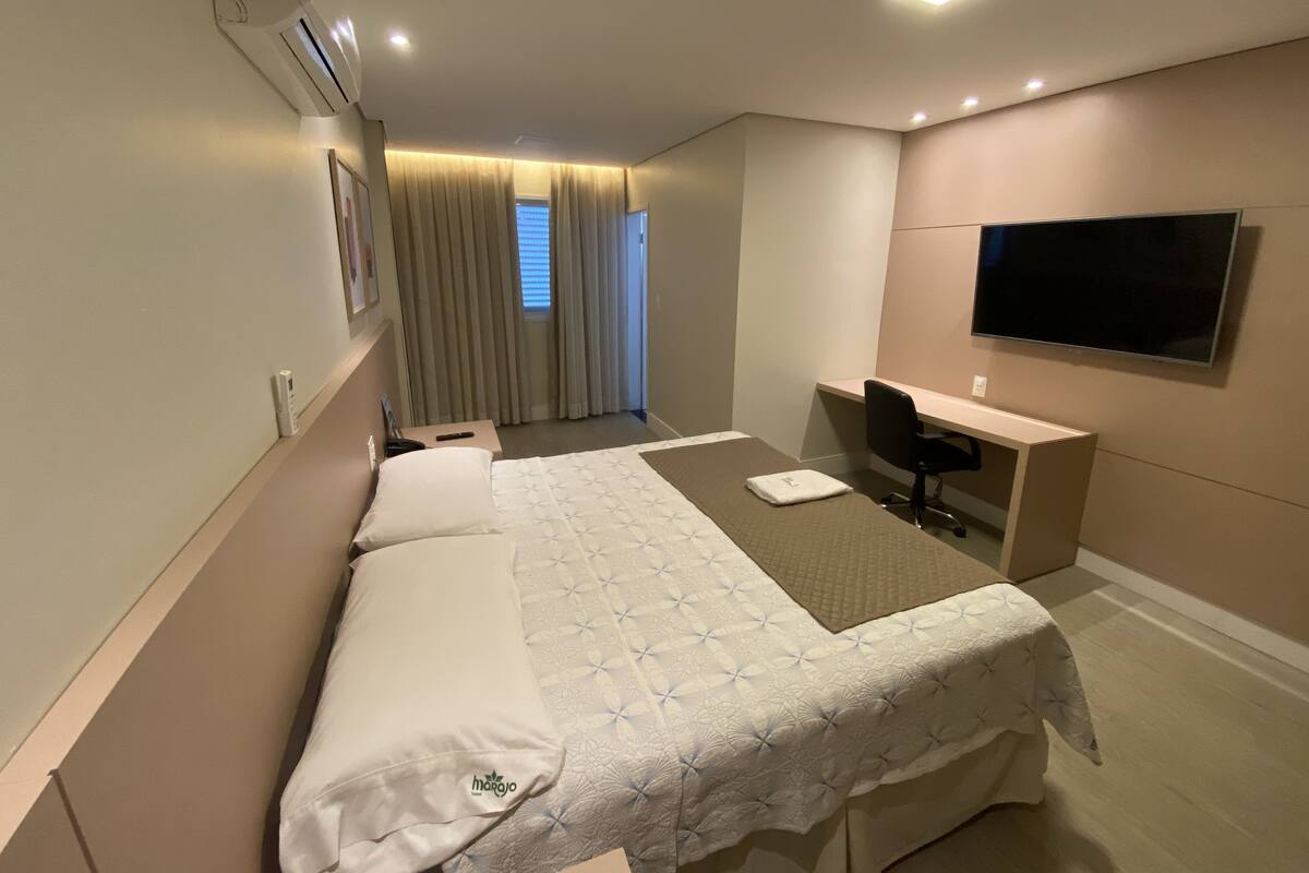 Quarto de hotel com cama, escrivaninha, cadeira, televisão e ar condicionado.