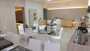 Un comedor moderno con sillas y mesas blancas, un mostrador de buffet con diferentes alimentos y una estación de café con un termo.