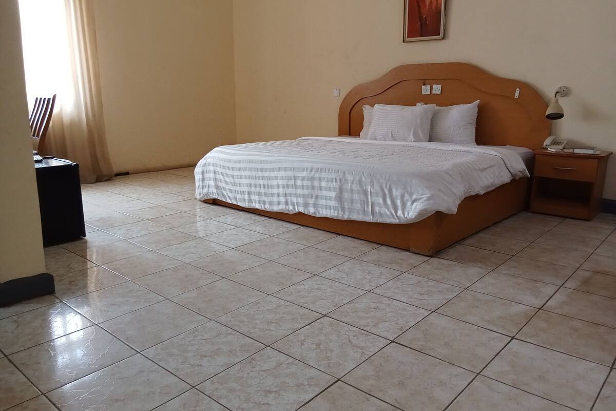 A neatly made bed with a white and gray checkered bedspread, a wooden headboard, and two white pillows. There is a small bedside table with a lamp and a telephone. The room has beige walls and a tiled floor.