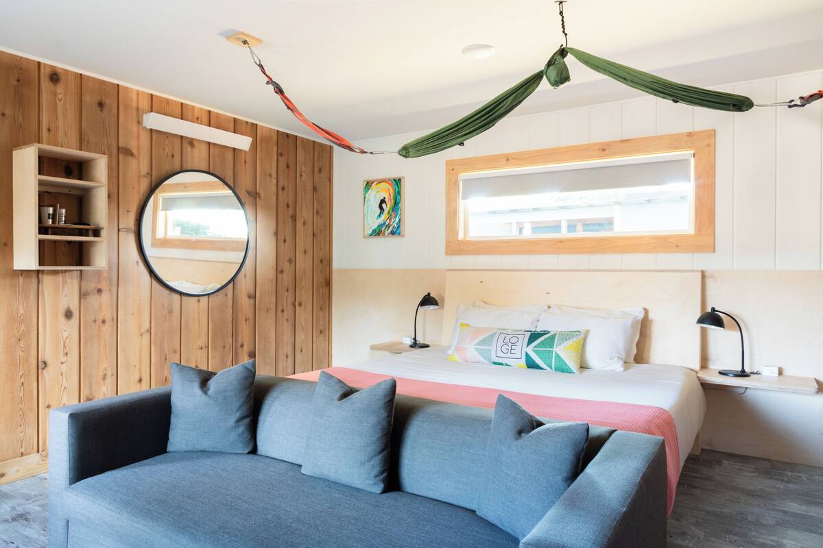 A room with a bed, a sofa, a hammock, a round mirror, and a wooden wall.