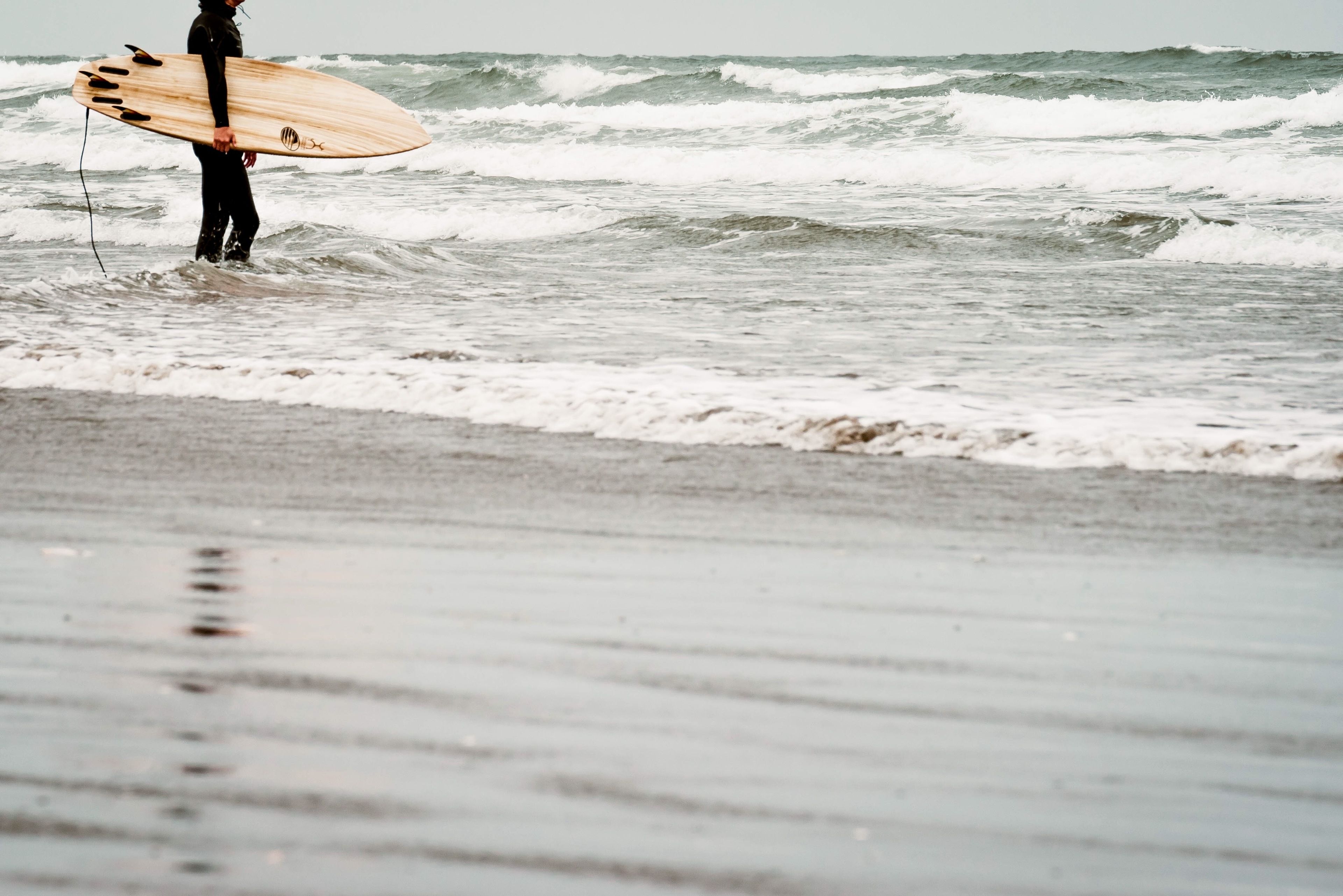 Eine Person in einem Neoprenanzug hält ein Surfbrett am Strand.