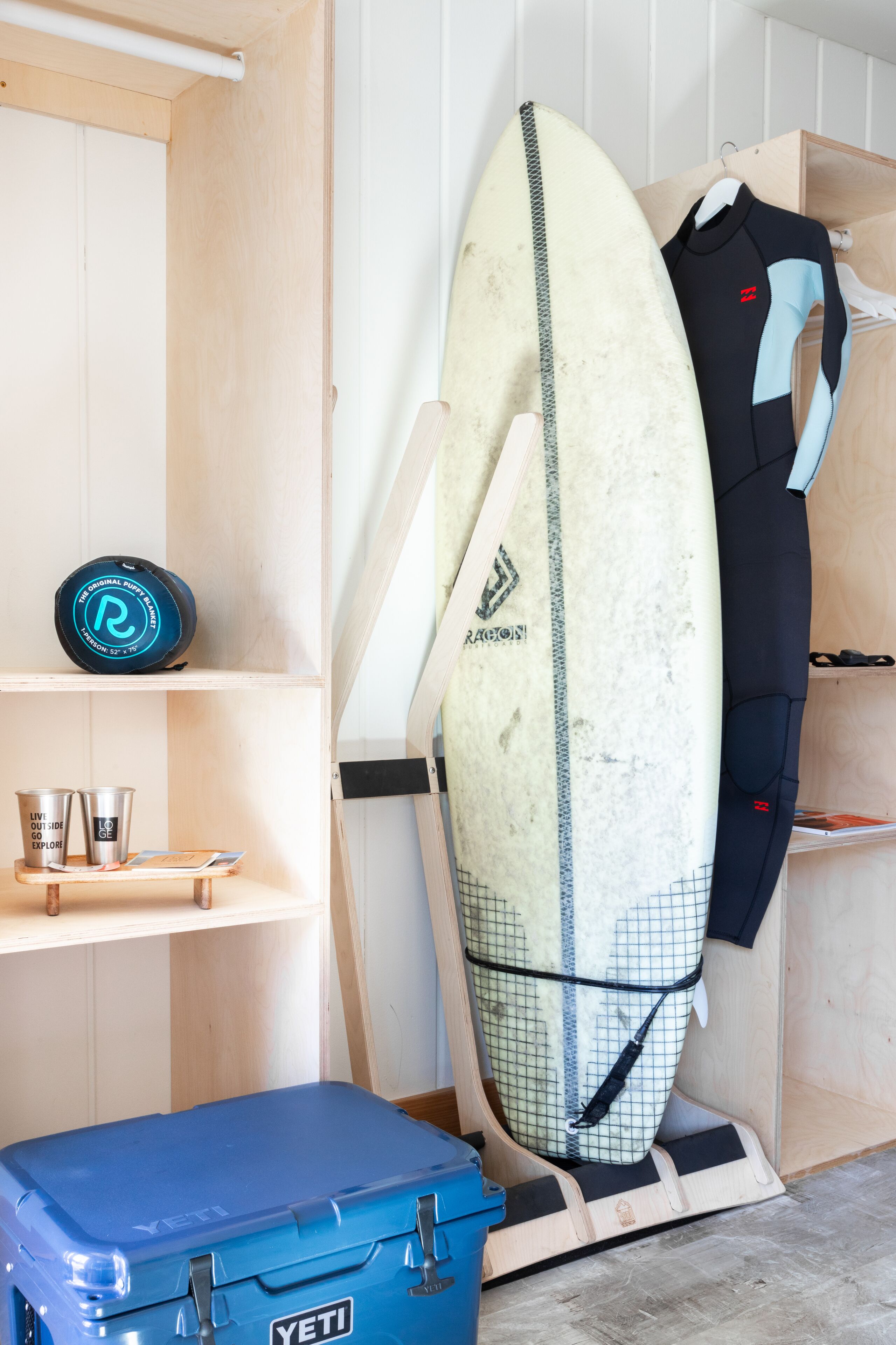 Une planche de surf appuyée contre un mur, une combinaison de plongée suspendue à un porte-vêtements et une boîte de rangement bleue étiquetée « YETI ».