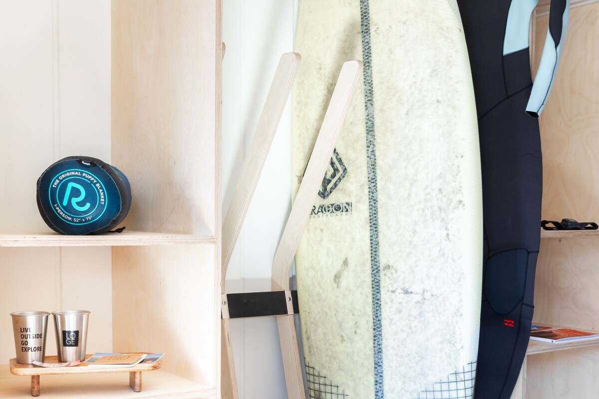 Ein Surfbrett, das an einer Wand lehnt, ein Neoprenanzug, der an einem Gestell hängt, und eine blaue Aufbewahrungsbox mit dem Aufdruck "YETI".