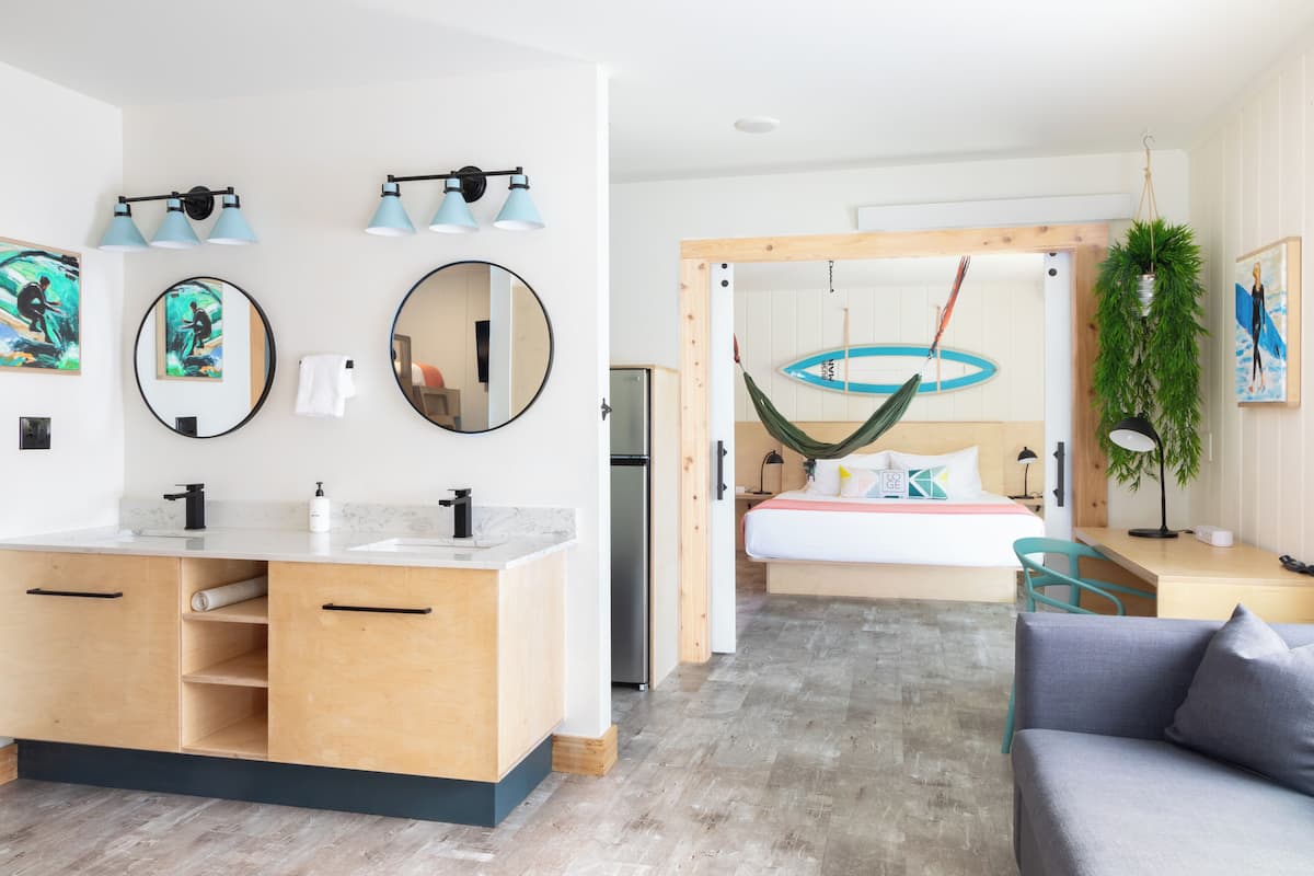 A modern hotel room with a large bathroom, a bed with a hammock, a desk, and a sofa.