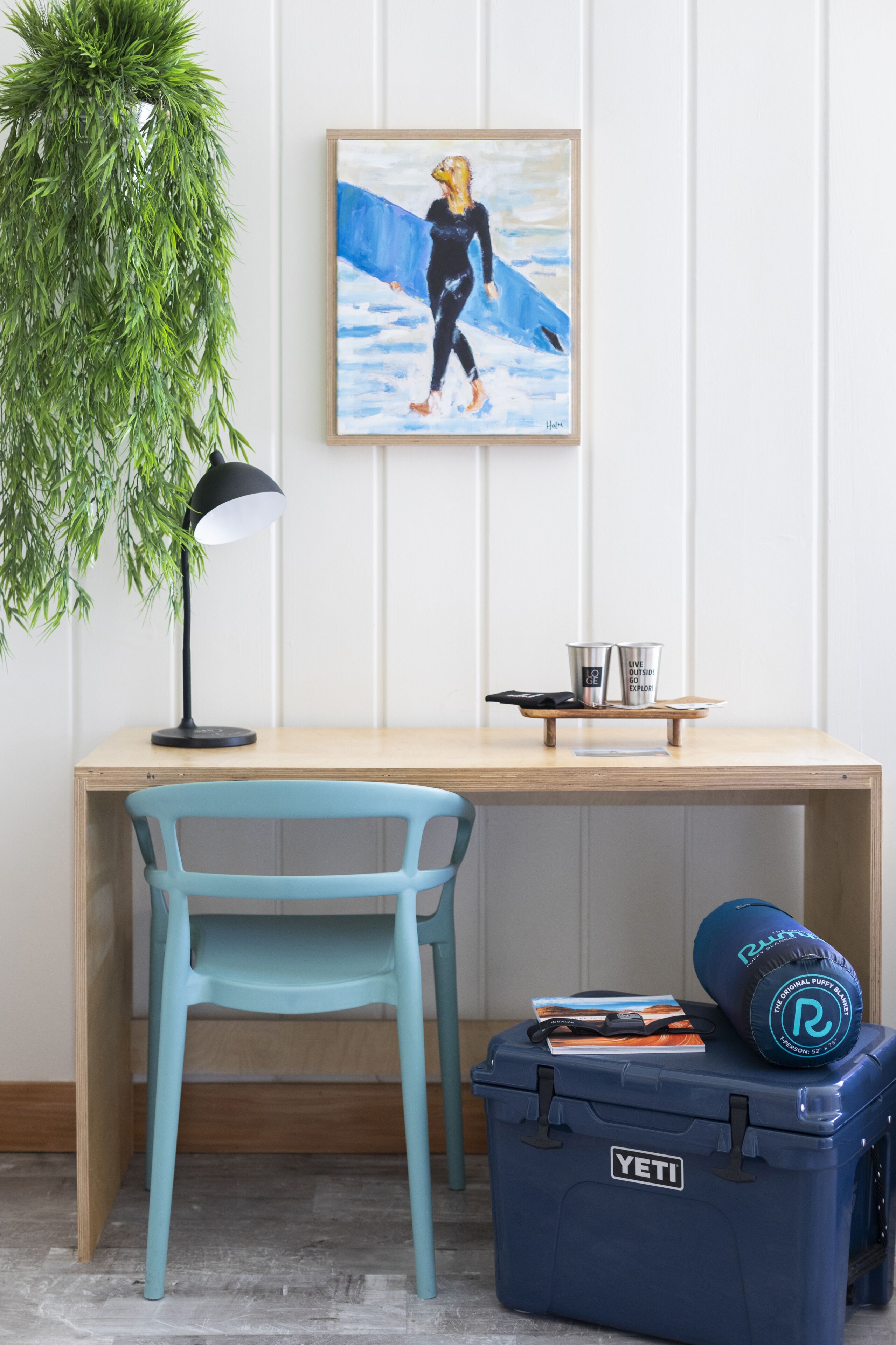 Un bureau avec une chaise bleue, un tableau encadré, une lampe de bureau et une boîte de rangement bleue contenant une planche de surf.