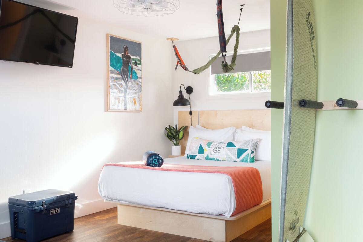A bedroom with a bed, a TV, a painting, a plant, and a suitcase.