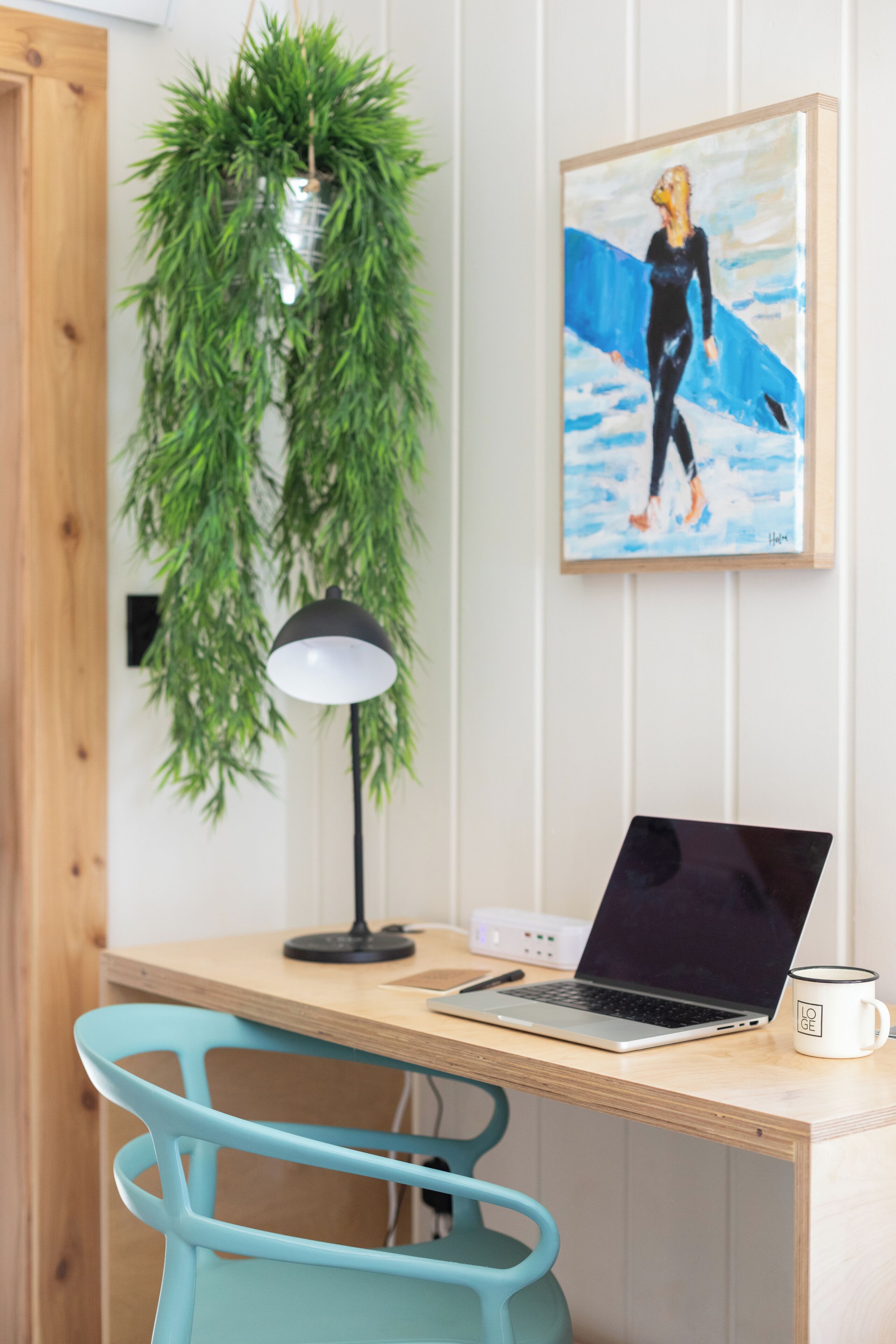 A workspace with a laptop, a mug, a desk lamp, a hanging plant, and a framed painting.